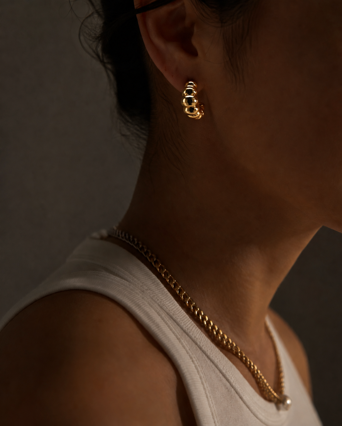 A close-up lifestyle shot of a person wearing a gold huggie hoop earring and a gold chain necklace, shown in warm, low-key lighting against a dark gray background.