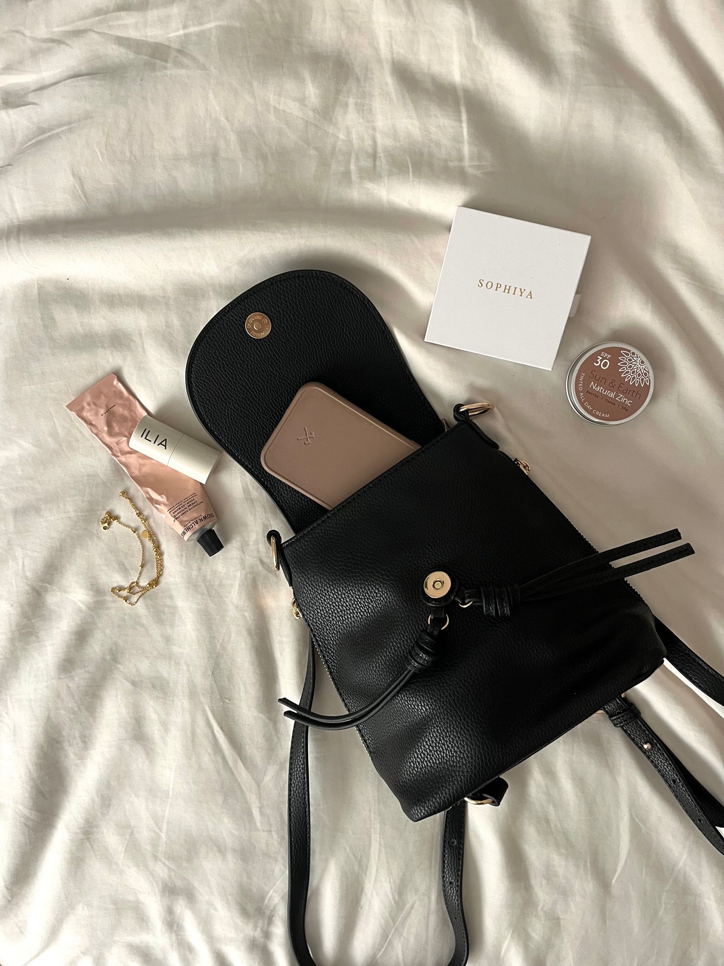 A neatly arranged flat lay showing the compact essentials that fit into a small shoulder bag. Items include a slim wallet, phone, keys, travel-size hand sanitizer, lip balm and sunglasses.