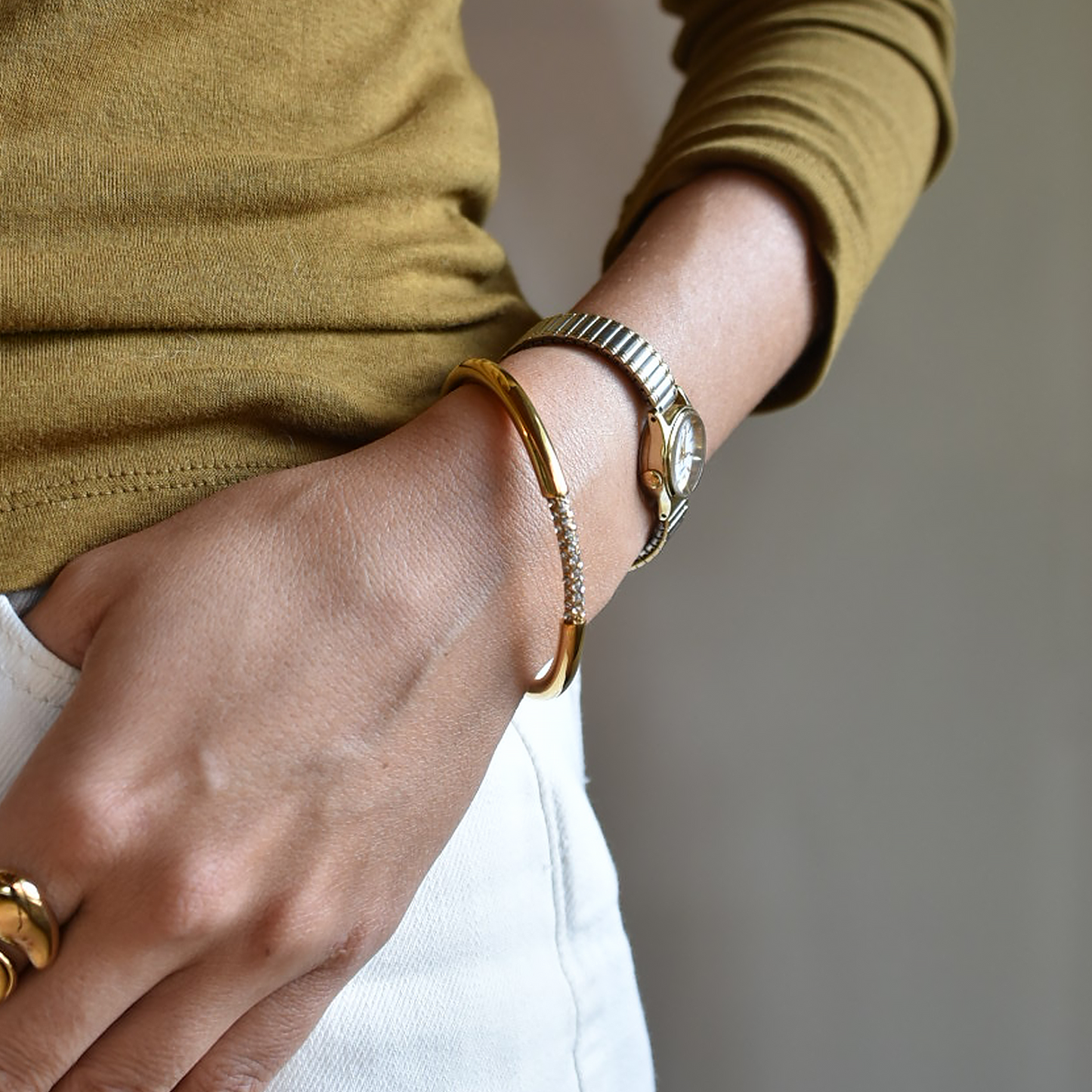 A close-up of a person's wrist wearing a polished gold bangle bracelet and a silver-and-gold watch, paired with a mustard yellow long-sleeve top and white pants.