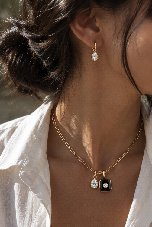 A close-up lifestyle shot of a model wearing a gold chain necklace with a double pendant design: a teardrop-shaped clear stone and a small black shield with a clear stone, paired with a matching teardrop earring.
