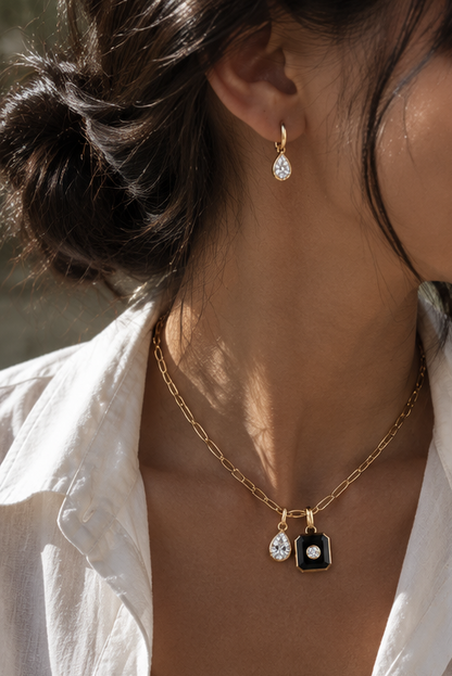 A close-up lifestyle shot of a model wearing a gold chain necklace with a double pendant design: a teardrop-shaped clear stone and a small black shield with a clear stone, paired with a matching teardrop earring.