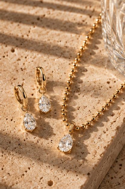 Gold jewelry set featuring a teardrop pendant necklace and matching drop earrings with clear teardrop gemstones, arranged on a textured beige stone surface with warm sunlight and shadows.