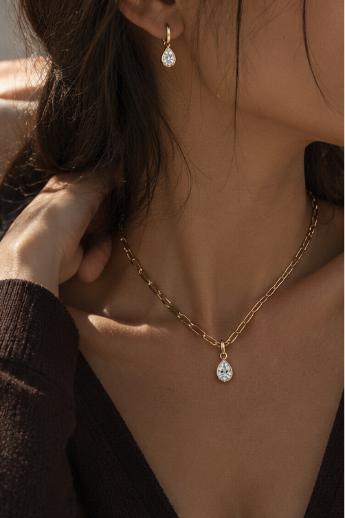 A close-up lifestyle shot of a model wearing a delicate gold chain necklace with a teardrop-shaped clear gemstone pendant, matching gold drop earrings, and a dark brown knit top.