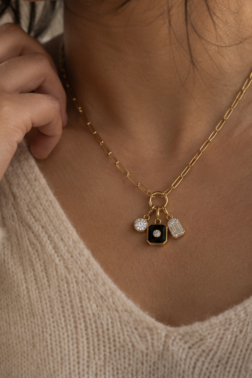 A close-up of a model wearing a gold chain necklace with a black square charm featuring a white center, flanked by two small clear crystal pendants, styled with a cream knit sweater.