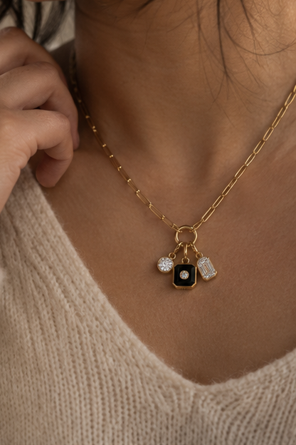 A close-up of a model wearing a gold chain necklace with a black square charm featuring a white center, flanked by two small clear crystal pendants, styled with a cream knit sweater.