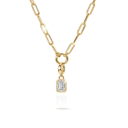 A gold paperclip chain necklace with a small rectangular emerald-cut clear gemstone pendant, shown on a clean white background with a soft reflection below.