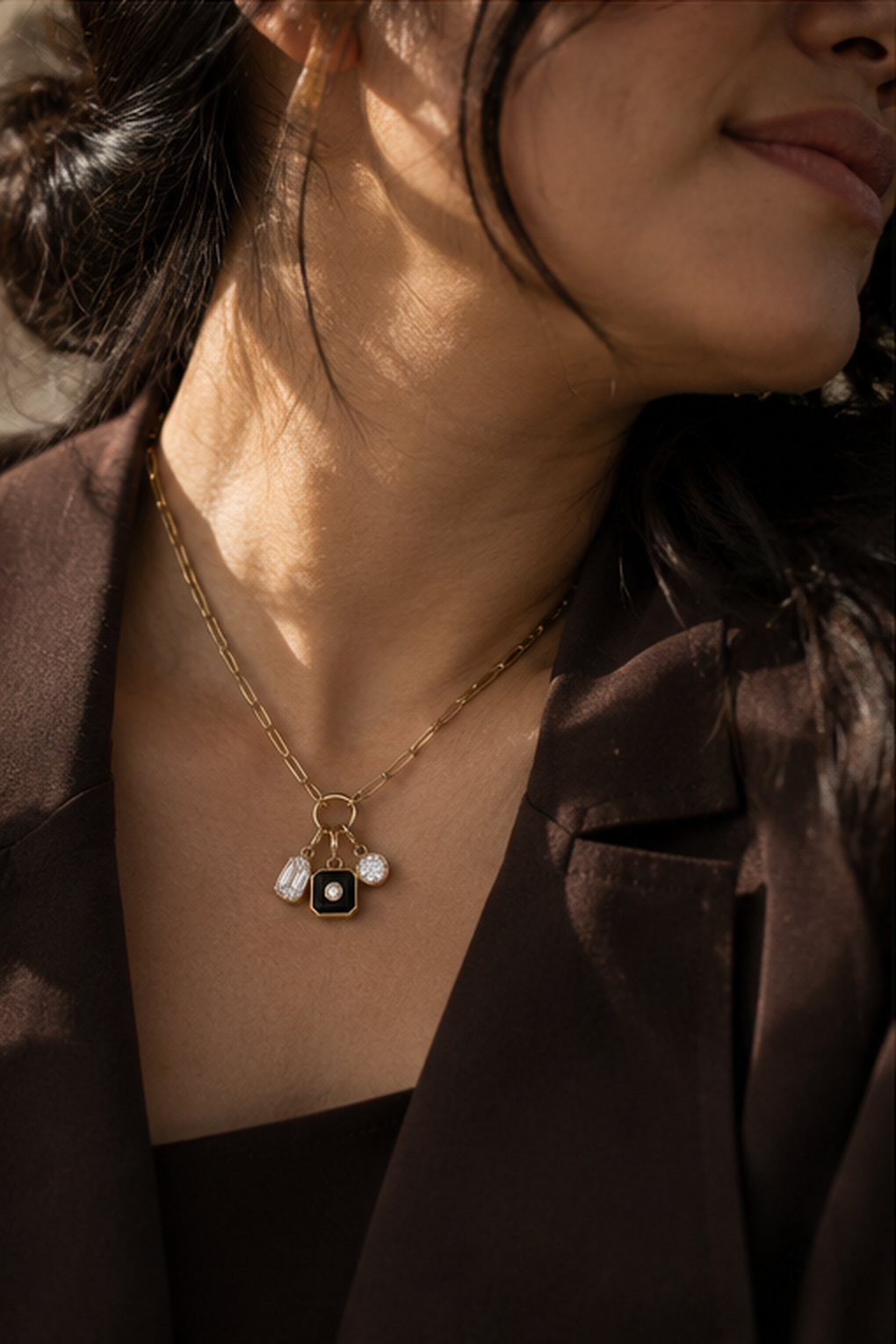 A close-up lifestyle shot of a woman wearing a delicate gold chain necklace with a central black square pendant and small white flower charms, styled with a dark brown blazer.