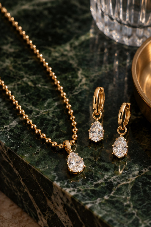 Gold jewelry set featuring a beaded chain necklace and matching drop earrings with pear-cut clear gemstones, displayed on a dark green marble surface.