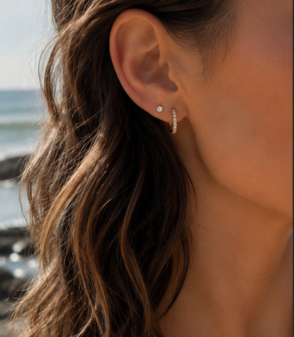 A close-up of a woman's ear wearing a small gold huggie hoop earring with tiny clear stones and a small silver stud earring, with her hair and a blurred outdoor background visible.