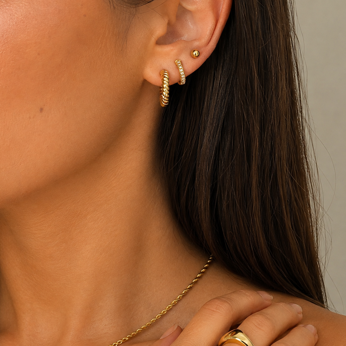 Close-up of a model's ear and neck wearing gold jewelry, including a twisted hoop earring, a small stud, a chain necklace, and rings on the hand.