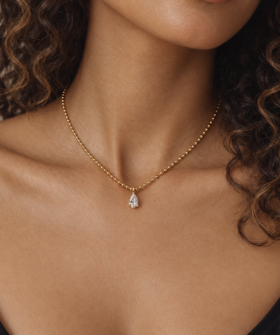 A close-up of a model wearing a delicate gold chain necklace with a pear-cut clear gemstone pendant, styled with curly brown hair and a black top.