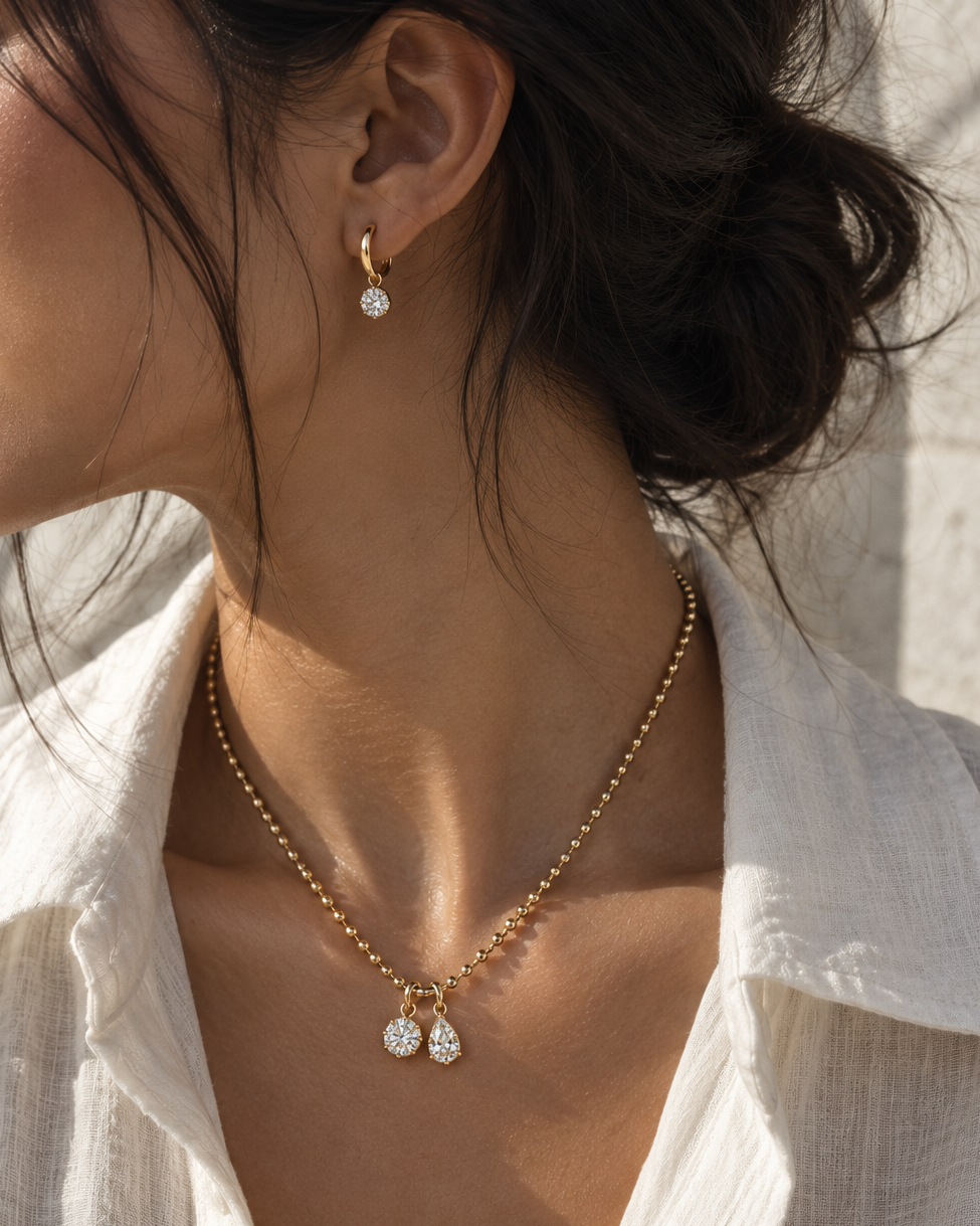 A close-up lifestyle shot of a woman wearing a delicate gold necklace with two small teardrop-shaped clear gemstone pendants, styled with a matching gold huggie hoop earring.