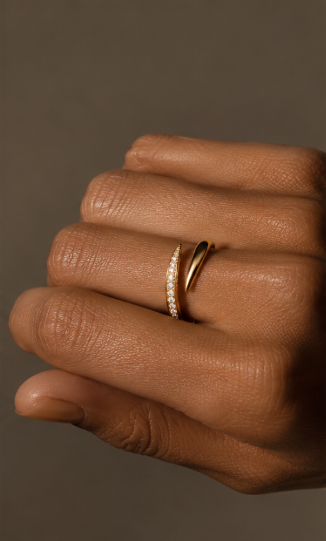 A close-up of a hand wearing two slim gold rings against a warm taupe background. One ring features a curved row of small clear stones, and the other is a simple polished gold band.