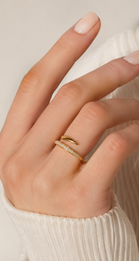 A close-up of a hand wearing two delicate gold rings, one with a small pavé band and another with a simple curved accent, paired with a cream ribbed sweater.