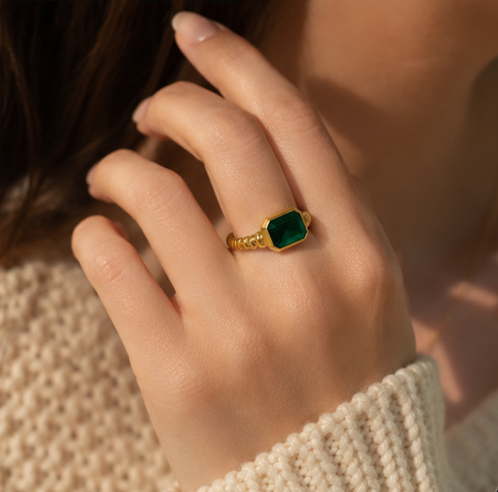 A close-up lifestyle shot of a person's hand wearing a gold ring with a green emerald-cut stone, set against a warm, softly blurred background.