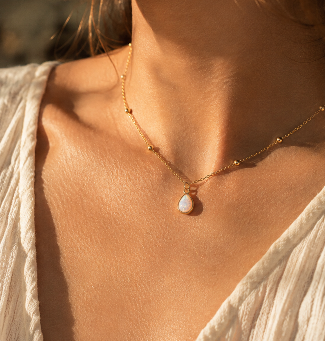 A close-up lifestyle shot of a woman wearing a delicate gold chain necklace with a small white teardrop pendant, set against warm golden-hour sunlight.