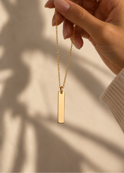 A gold bar pendant necklace held by a hand against a warm beige background with soft leaf shadows.