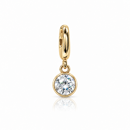 A gold huggie hoop earring with a round bezel-set clear gemstone, shown on a clean white background with a soft reflection below.