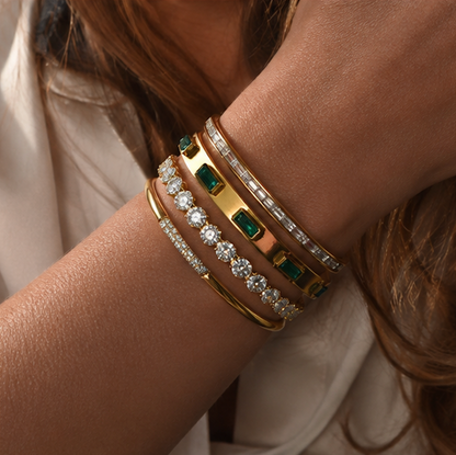 A close-up of a person's wrist wearing five stacked bangle bracelets. The bracelets include yellow gold, white gold, and silver-toned metal bands, some decorated with clear pavé diamonds and others with rectangular green emerald-cut stones.