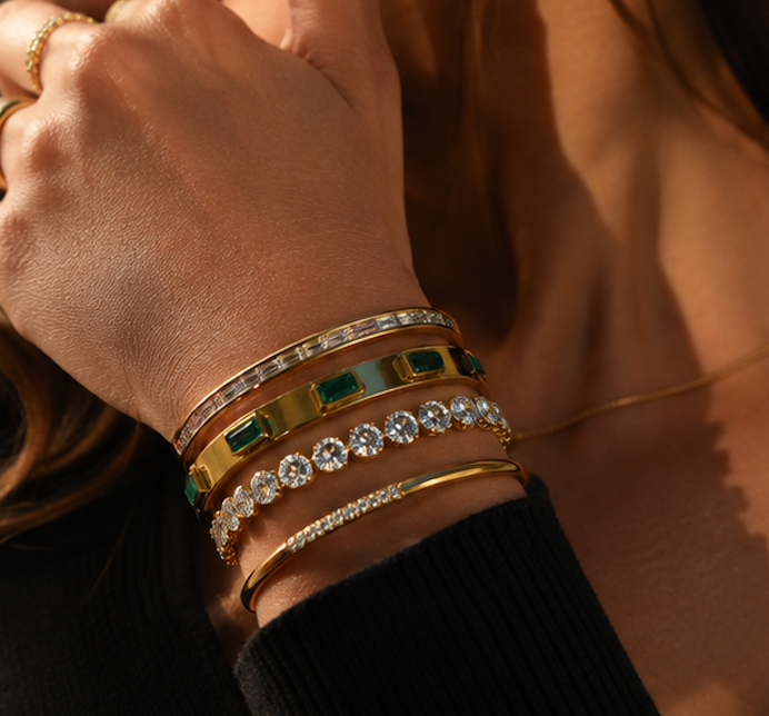 A close-up of a person's wrist wearing five stacked gold bangles, including two with emerald-cut green stones, two with clear round stones, and one with small clear stones, against warm golden lighting.