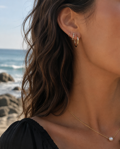 A close-up of a woman's ear wearing a stack of gold hoop and stud earrings, with a gold chain necklace and a pearl earring, set against a blurred beach and ocean background.