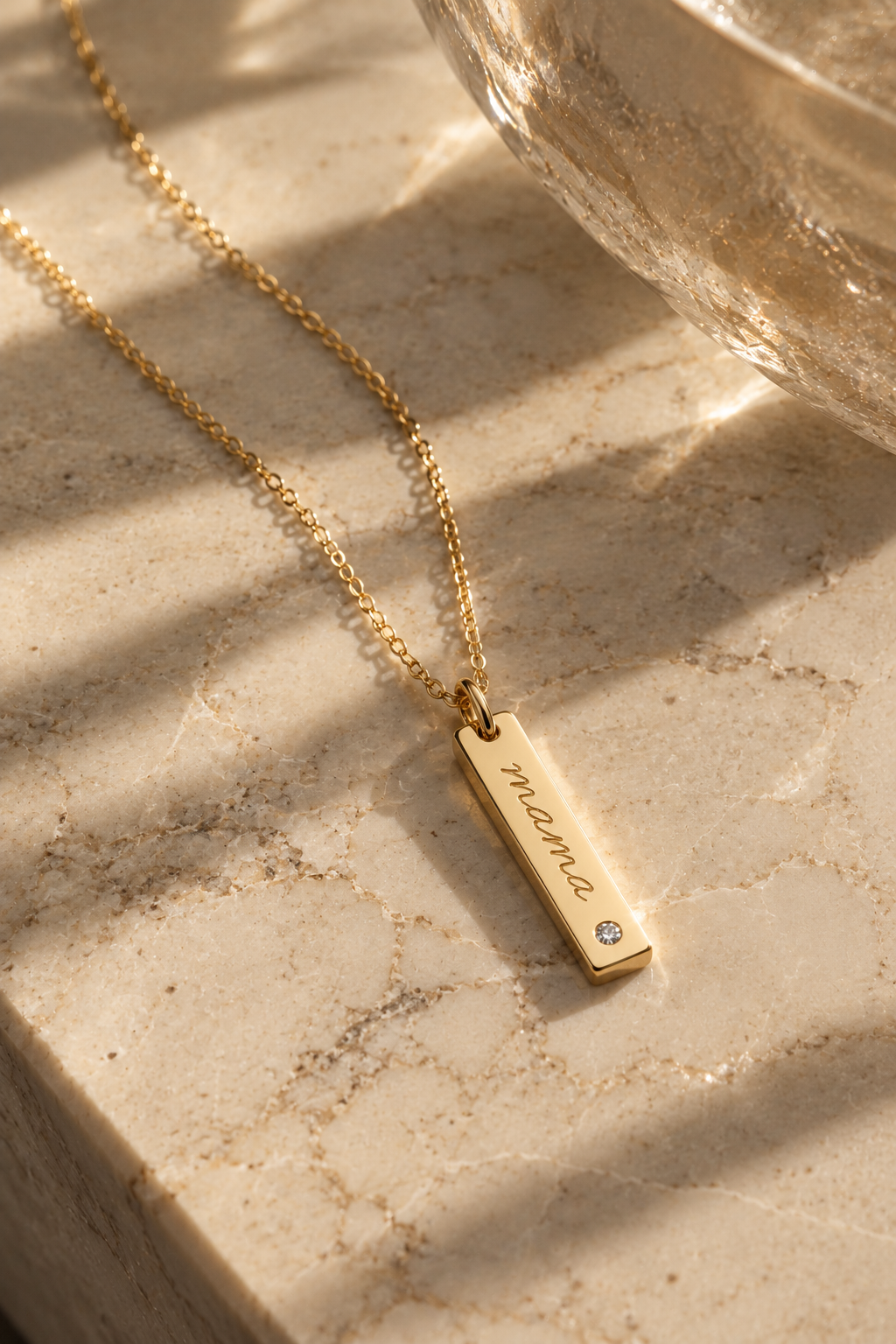 A gold bar pendant necklace engraved with the name 'Mia' and a small clear gemstone, resting on a beige marble surface with warm sunlight and shadows.