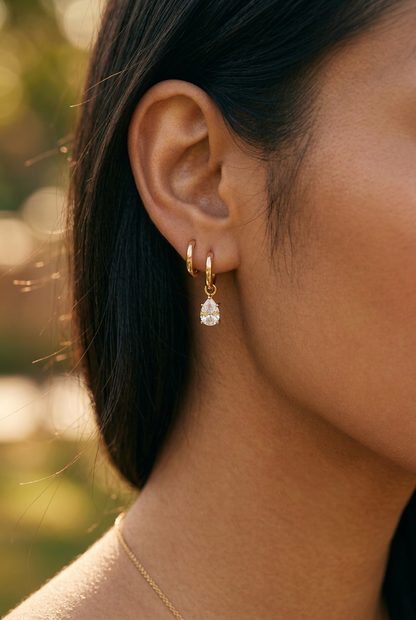 A close-up of a woman's ear wearing a delicate gold huggie hoop earring with a dangling teardrop-shaped clear gemstone.