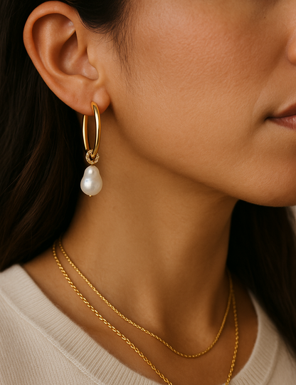 A close-up of a woman wearing a gold hoop earring with a dangling white pearl, layered with two delicate gold necklaces, against a warm neutral background.