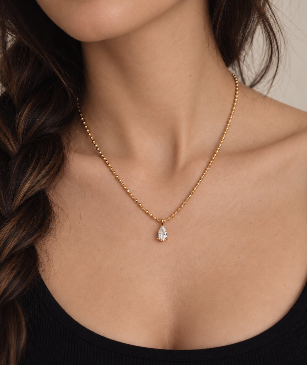 A close-up of a woman wearing a delicate gold chain necklace with a small teardrop-shaped clear gemstone pendant, styled with a black ribbed top against a neutral beige background.