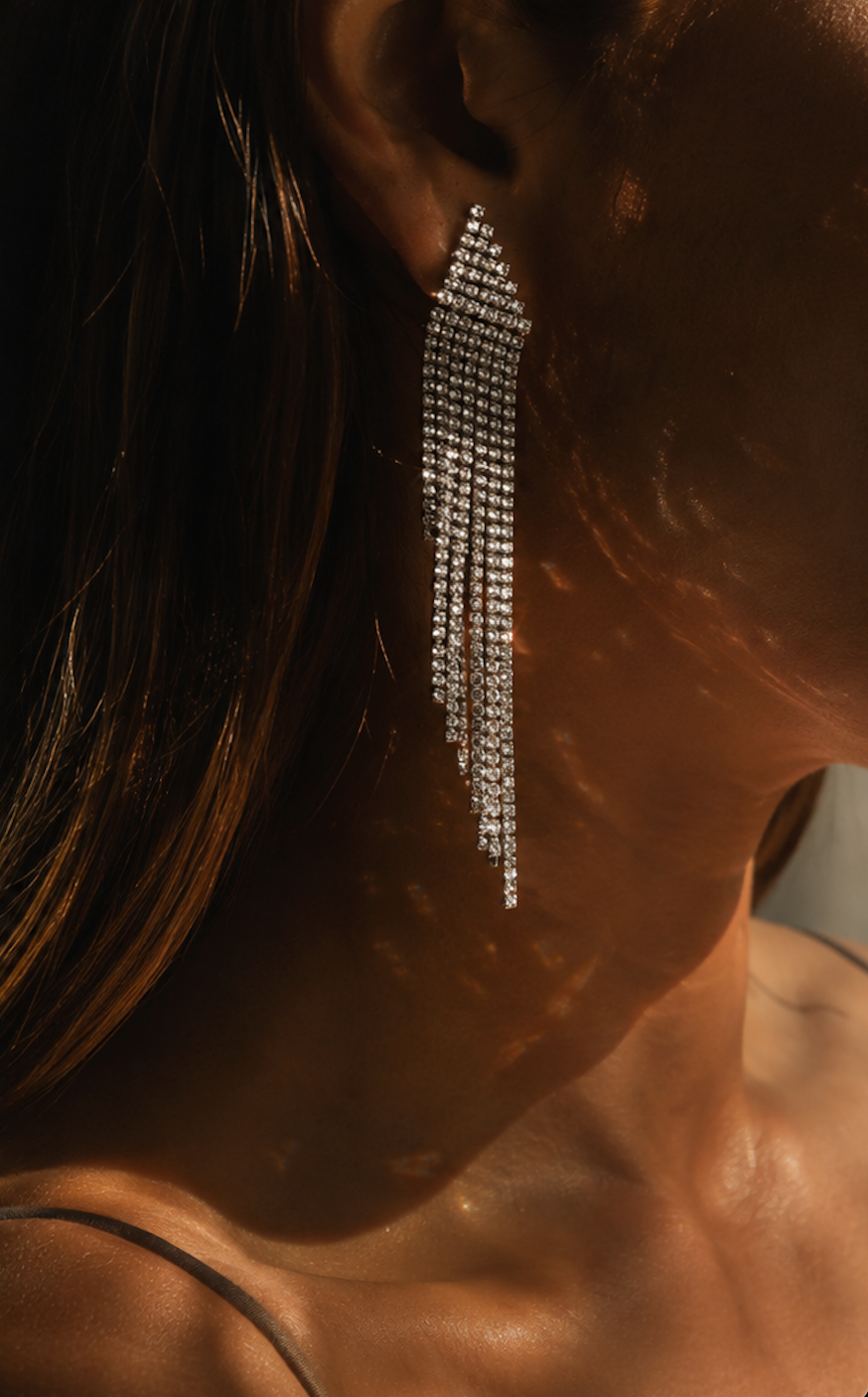 A close-up lifestyle shot of a model wearing a long, dangling statement earring made of sparkling clear stones in a silver-tone setting. The warm, dramatic lighting casts strong shadows on the model's skin and hair.