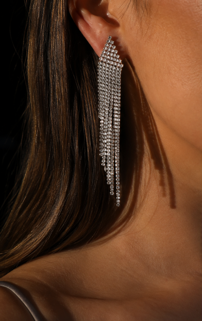 A close-up of a woman's ear wearing a long, sparkling silver dangle earring with multiple rows of clear rhinestones. Her brown hair and skin are visible against a dark background.
