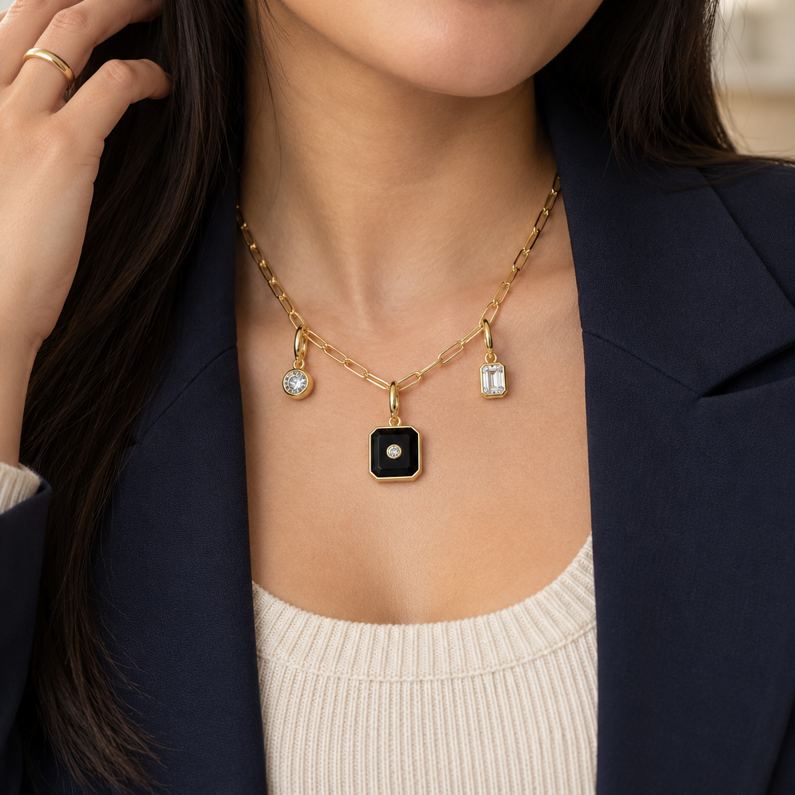 A close-up of a woman wearing a gold chain necklace with a black square pendant featuring a white center, along with two small clear stone charms. She is wearing a navy blazer and a cream ribbed top.