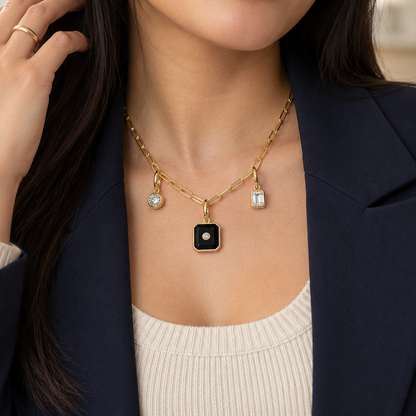 A close-up of a woman wearing a gold chain necklace with a black square pendant featuring a white center, along with two small clear stone charms. She is wearing a navy blazer and a cream ribbed top.
