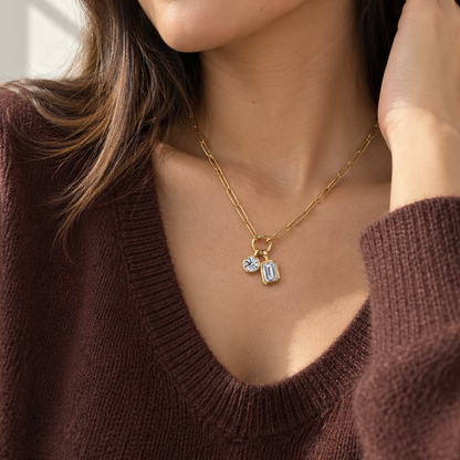 A close-up lifestyle shot of a woman wearing a delicate gold chain necklace with two small clear gemstone charms, paired with a brown knit sweater.