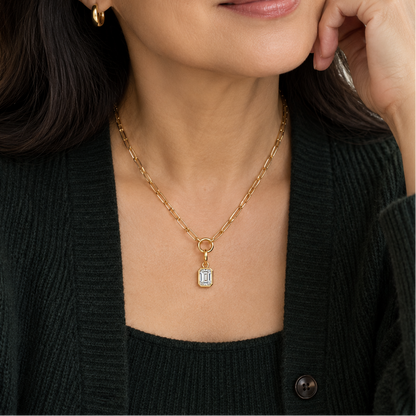 A close-up of a woman wearing a delicate gold chain necklace with a rectangular clear gemstone pendant, styled with a dark green cardigan.