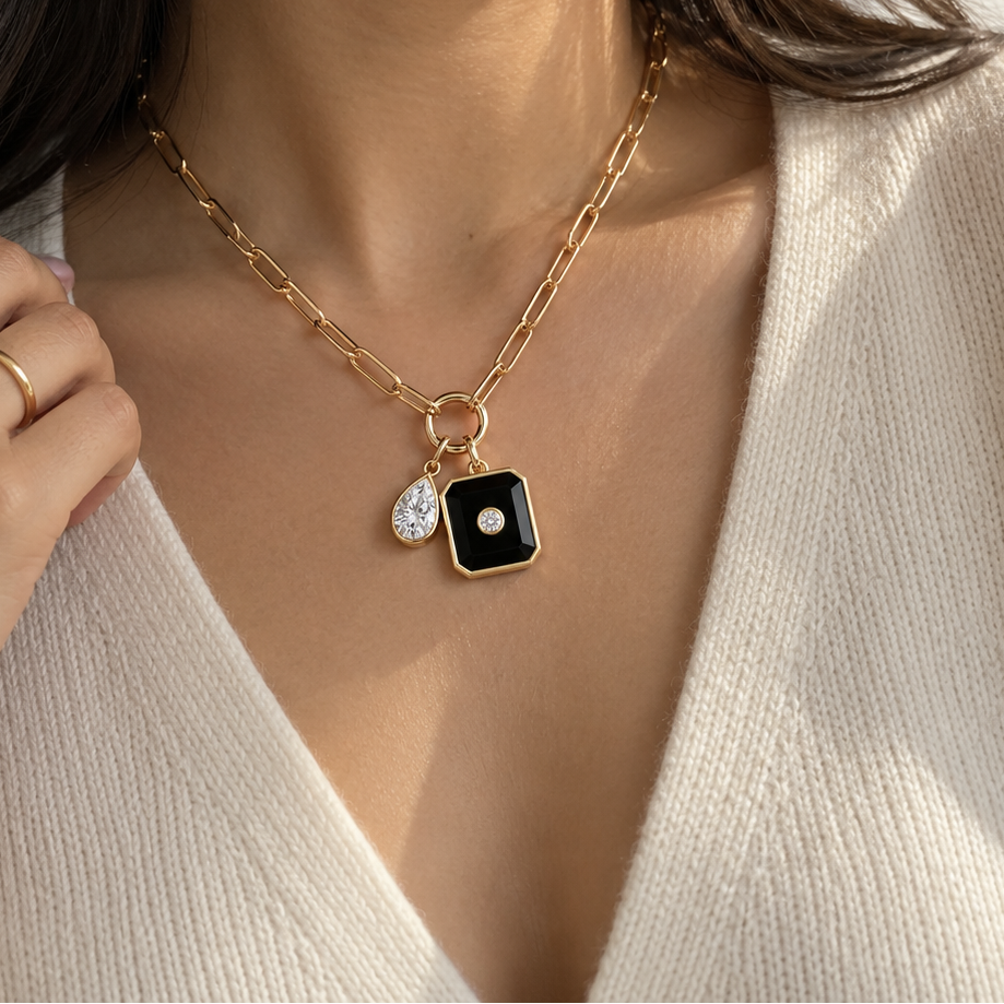 A close-up lifestyle shot of a model wearing a gold chain necklace with a black square pendant and a small teardrop-shaped clear stone charm.