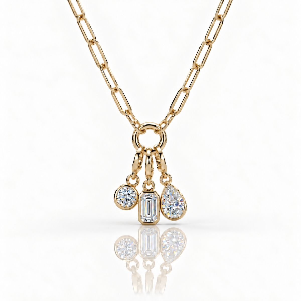 A gold chain necklace with a charm ring holding three dangling clear gemstones in round, rectangular, and teardrop shapes, shown on a clean white background with a soft reflection below.