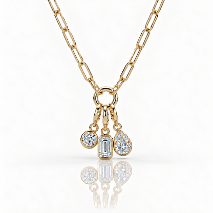A gold chain necklace with a charm ring holding three dangling clear gemstones in round, rectangular, and teardrop shapes, shown on a clean white background with a soft reflection below.