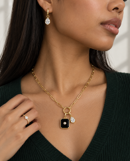 A close-up lifestyle shot of a model wearing a gold chain necklace with a black square pendant and a small teardrop charm, along with matching drop earrings and a ring.