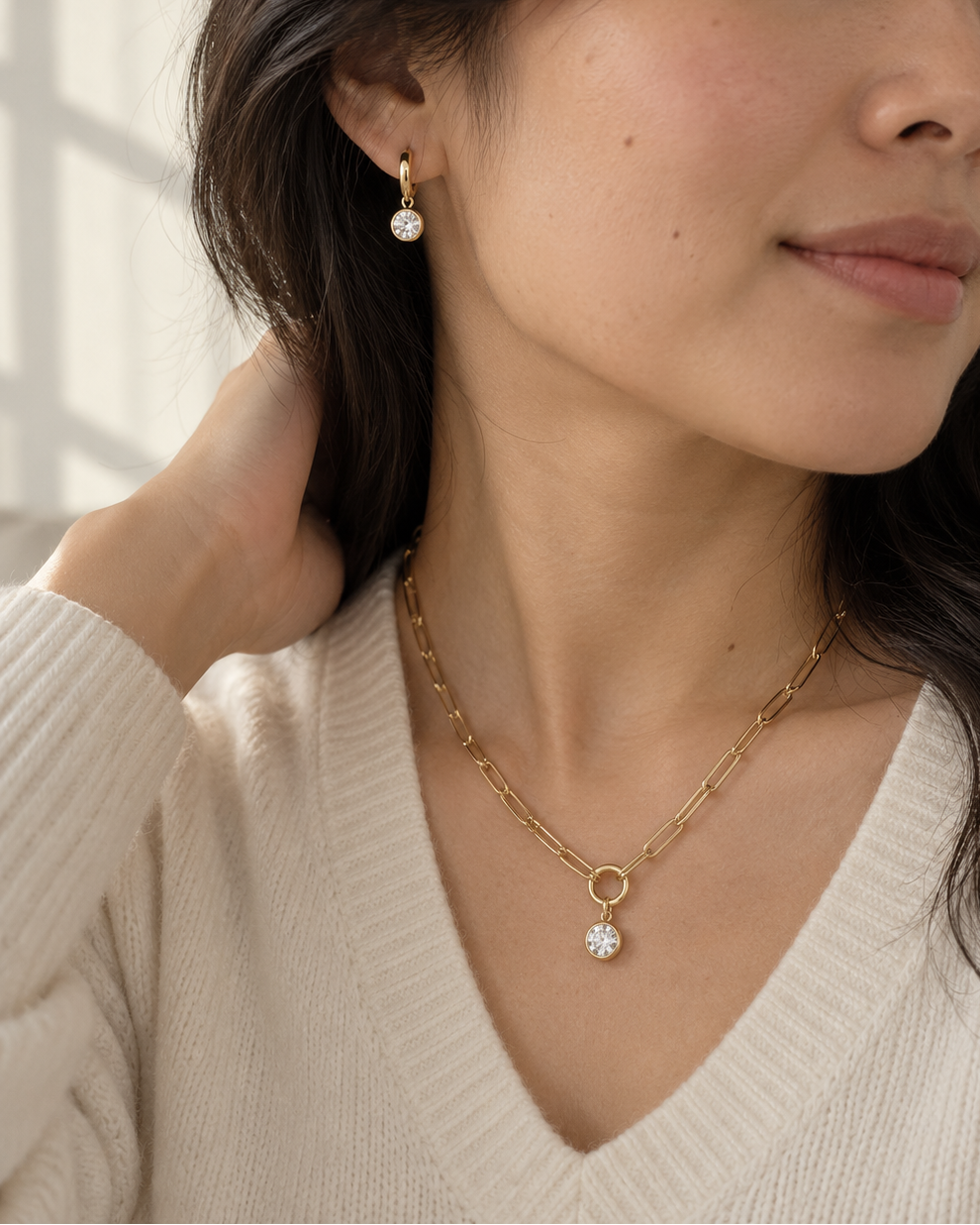 A close-up lifestyle shot of a woman wearing a delicate gold chain necklace with a small clear crystal pendant and a matching gold drop earring.