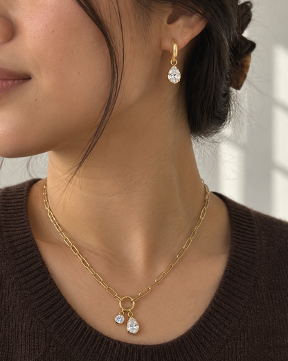 A close-up of a woman wearing a delicate gold chain necklace with two small teardrop-shaped clear crystal pendants and a matching gold drop earring.