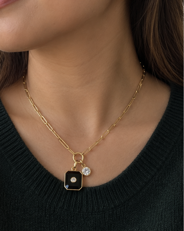 A woman wearing a delicate gold chain necklace with a black square pendant and a small clear stone, along with a matching drop earring, shown in a close-up portrait.