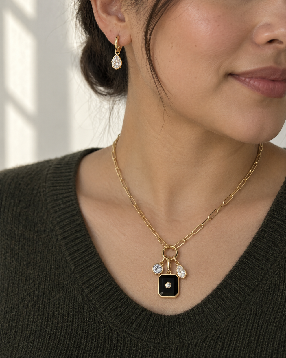 A woman wearing a gold chain necklace with a black rectangular pendant and small clear stones, paired with a matching gold drop earring, shown in a softly lit indoor setting.