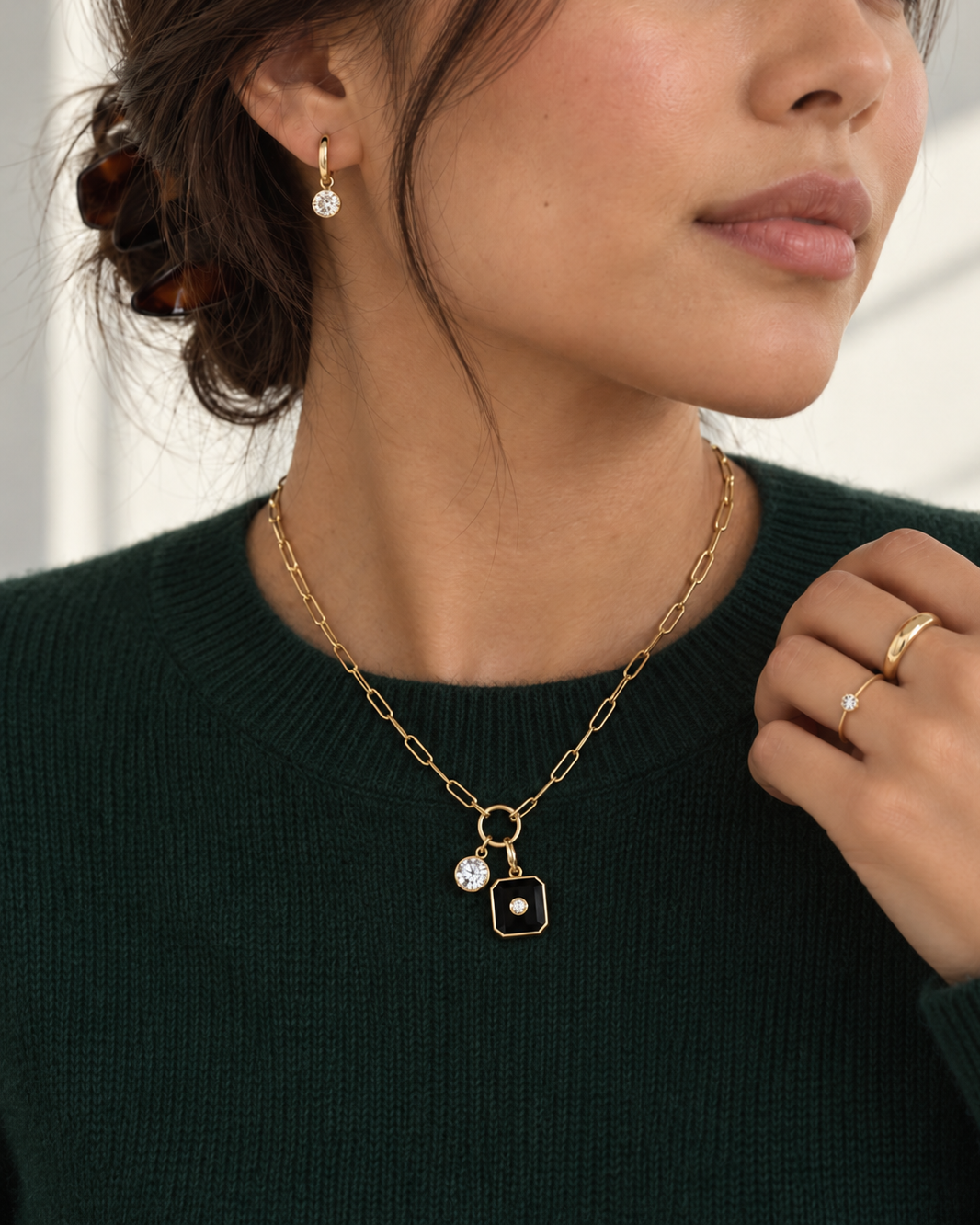 A close-up lifestyle shot of a woman wearing a gold chain necklace with a square pendant featuring a black center and a clear stone, along with matching gold hoop earrings and a ring.