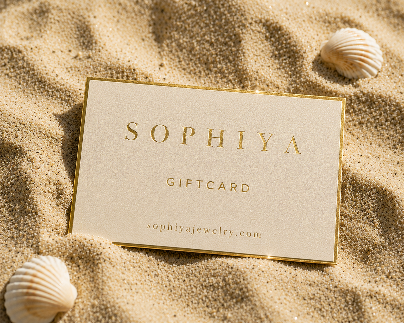 A rectangular Sophiya gift card with a gold border and gold foil lettering, placed on sand with seashells around it. The card reads 'SOPHIYA', 'GIFTCARD', and 'sophiyajewelry.