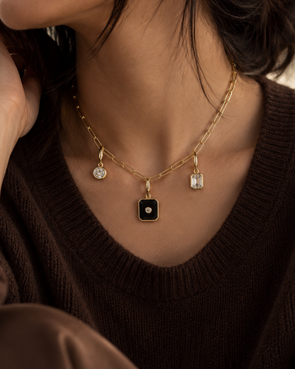 A close-up lifestyle shot of a model wearing a delicate gold chain necklace with a central square black pendant and two small clear crystal charms, styled with a dark brown knit sweater.