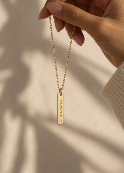 A gold bar necklace with engraved text and a small clear stone, held by a hand against a warm beige background with soft leaf shadows.