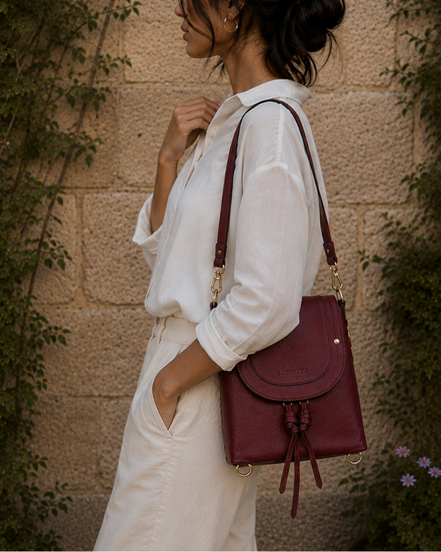 A woman in a white shirt and white pants carries a burgundy leather shoulder bag with gold hardware, standing against a stone wall with green vines.